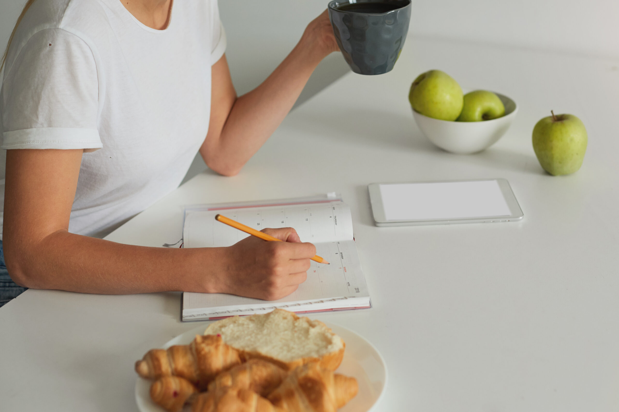 Close up woman hands, view from a side, planning her day, keeps Close up woman hands, view from a side, planning her day, keeps a grey cup with some liquid, Croissants on a plate, apples, phone with white screen for your text are there on a white kitchen table